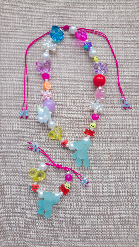 A children's beaded necklace and matching bracelet laid flat on a beige textured fabric background. The jewelry features a light blue elephant charm, white faux pearls, and colorful translucent beads in pink, blue, purple, yellow, and red.